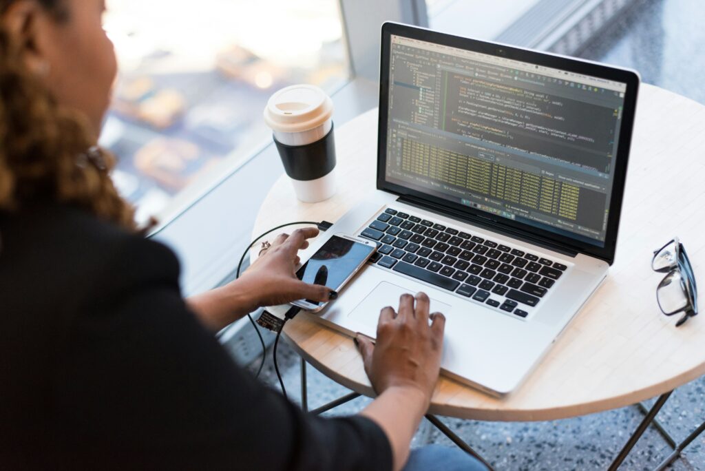 Person sitz mit ihrem Kaffee vor einem Laptop und hat ihr Handy in der Hand